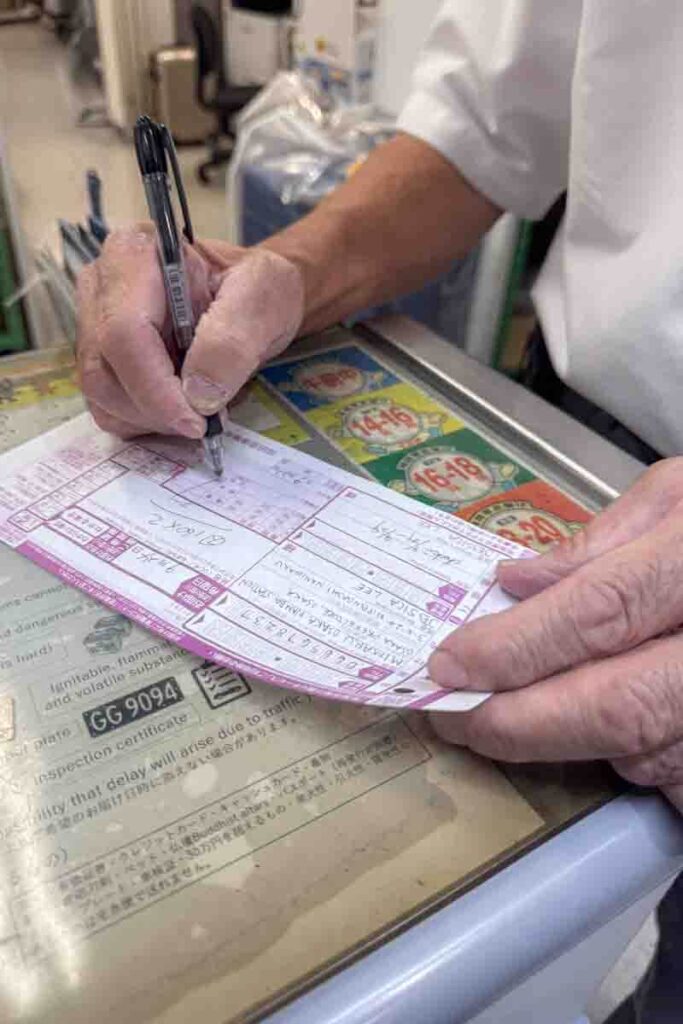 Hands of a Yamato staff member writing on a detailed shipping form at a desk covered in promotional stickers and instructions. Highlights the step-by-step care taken in the luggage forwarding process.