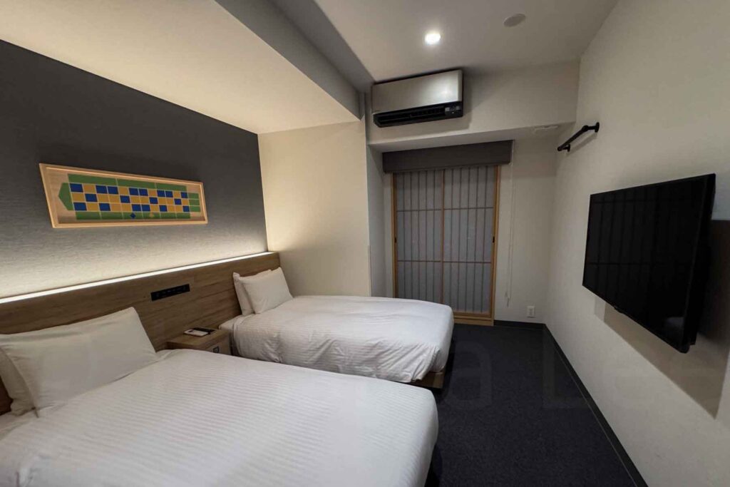 A snapshot of the main bedroom in the 2 bedroom Japanese apartment at Mimaru Osaka Namba Station, showcasing the clean layout and calm aesthetic that appeals to families with kids. The room has two single beds in crisp white linens, a low profile wood panel as a headboard, dark short carpet, and a shoji style sliding window shade. A TV is across from the beds and the floor as plenty of walking space around the beds.