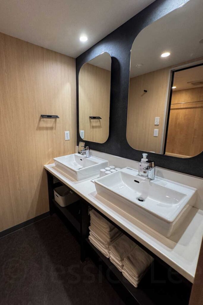 A spacious family bathroom at Mimaru Osaka Namba hotel featuring sleek Japanese style with wood tones, clean white features and linens and black accents showing dual square sinks, large mirrors, and stacked towels — ideal for sharing.