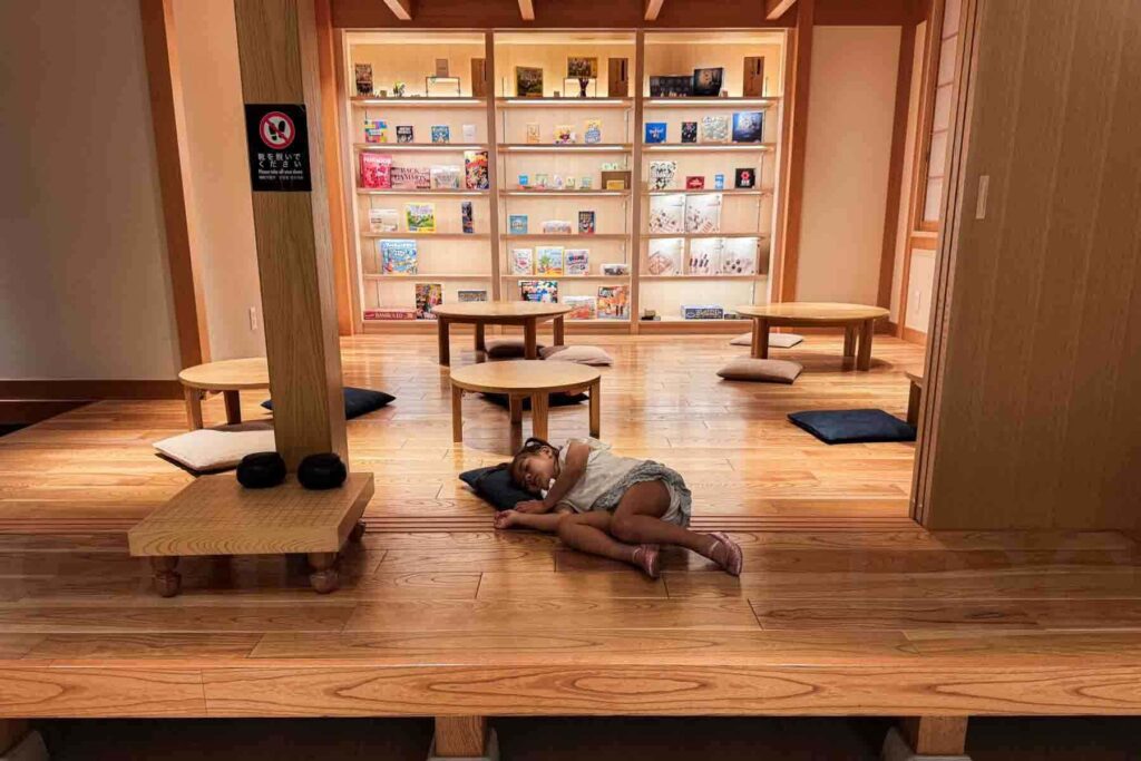A toddler sleeping on the raised Japanese wooden floor with her head on a pillow in front of board game shelves at the hotel’s play lounge in Osaka — a peaceful rest spot for kids.