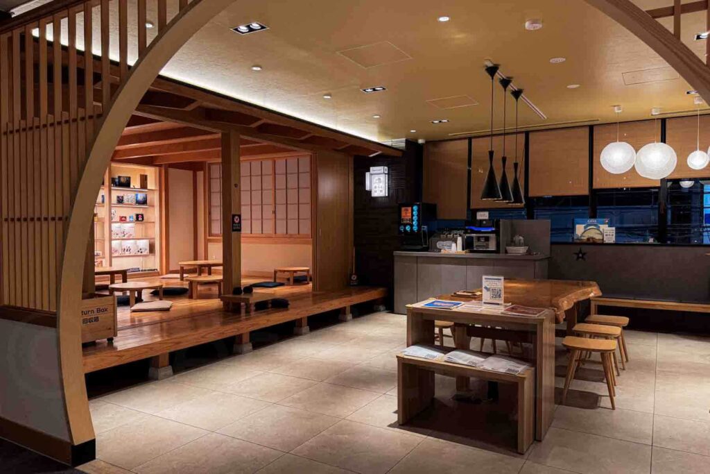 A warm lounge space with a raised wooden floor, decorative wooden arch, wooden bookshelves, and wooden tables, soft lighting, drink station and seating designed for families at a family-friendly apartment hotel in Osaka.