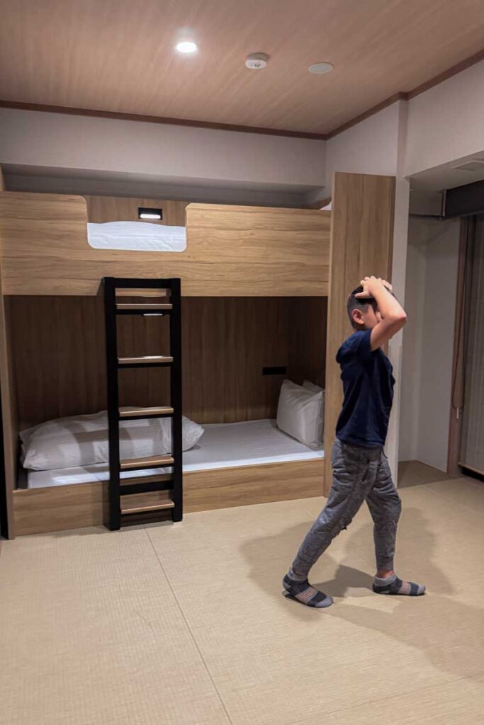 A family apartment room at the Mimaru Namba Station hotel in Osaka featuring warm wooden bunk beds, shown with a boy in a navy T-shirt and gray joggers enjoying the tatami mat room.