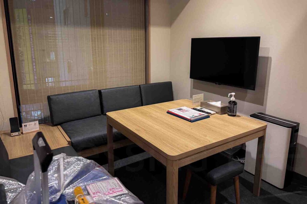 A combo living and dining area in a Mimaru hotel apartment in Osaka with a wooden table, TV and bench seating — great for family stays. There’s a large window behind the bench seat. The table features informational papers, tissues and hand sanitizer. A suitcase is barely visible in the bottom left corner.
