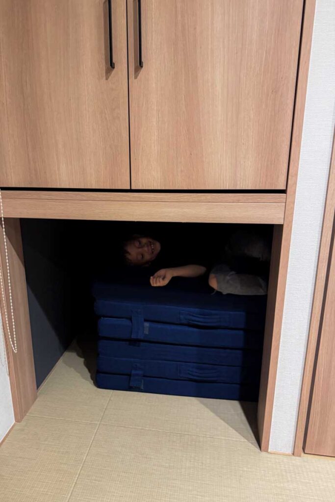 A child peeks out from beneath stacked blue futons in a cosy alcove inside a hotel room, under a raised wooden cabinet, demonstrating playful space for kids.