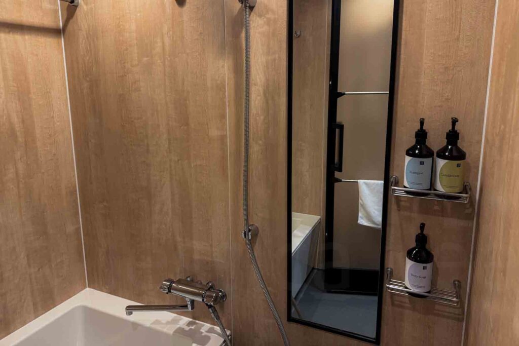 Close‑up of the shower wall with a handheld shower head, a full-length mirror, and wire shelves holding shampoo, conditioner and body soap in the hotel bathroom.