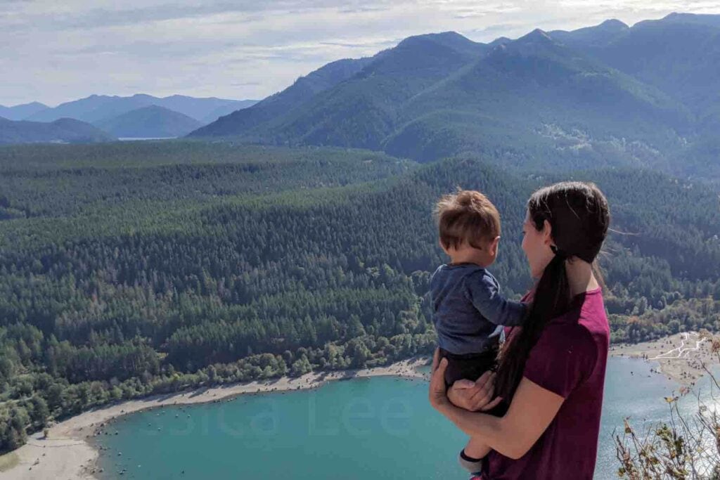 Mother holding her baby while standing on a rocky overlook high above Rattlesnake Lake with mountains stretching across the horizon. This scenic viewpoint shows why the trail is one of the most popular hikes near Seattle.