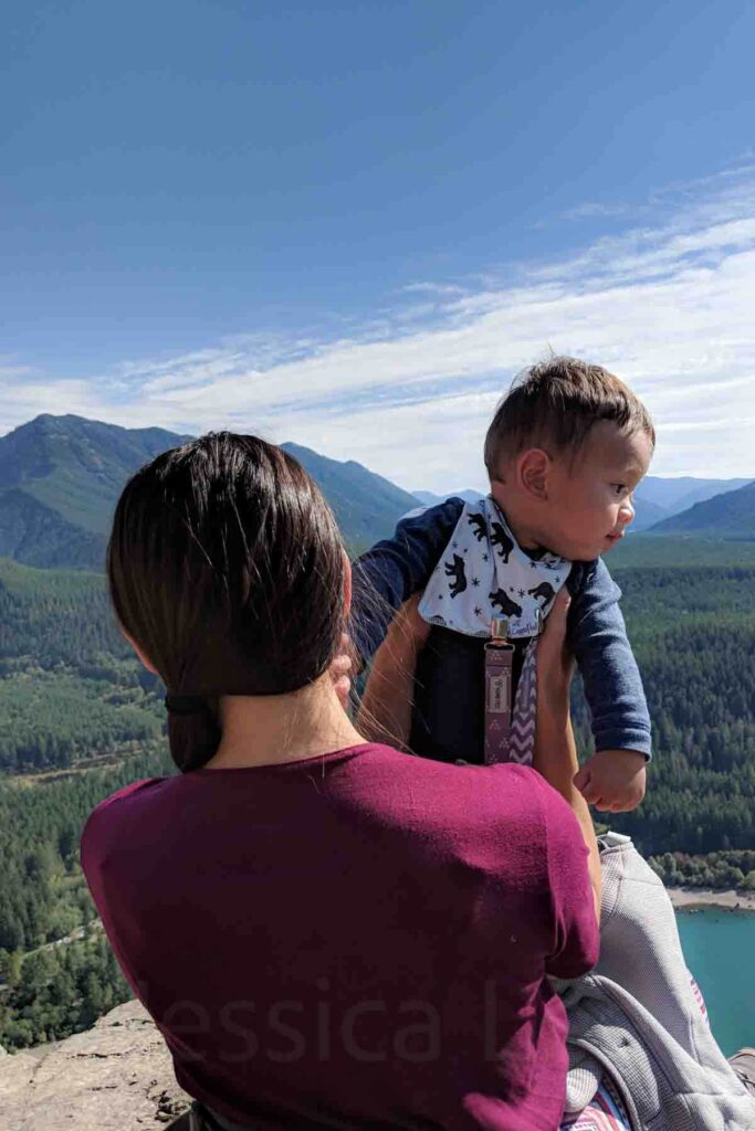 Mom holding a baby while looking out over the blue waters of Rattlesnake Lake and forest covered mountains from Rattlesnake Ledge. Scenic viewpoints like this are a highlight of hikes near Seattle and make the climb rewarding.