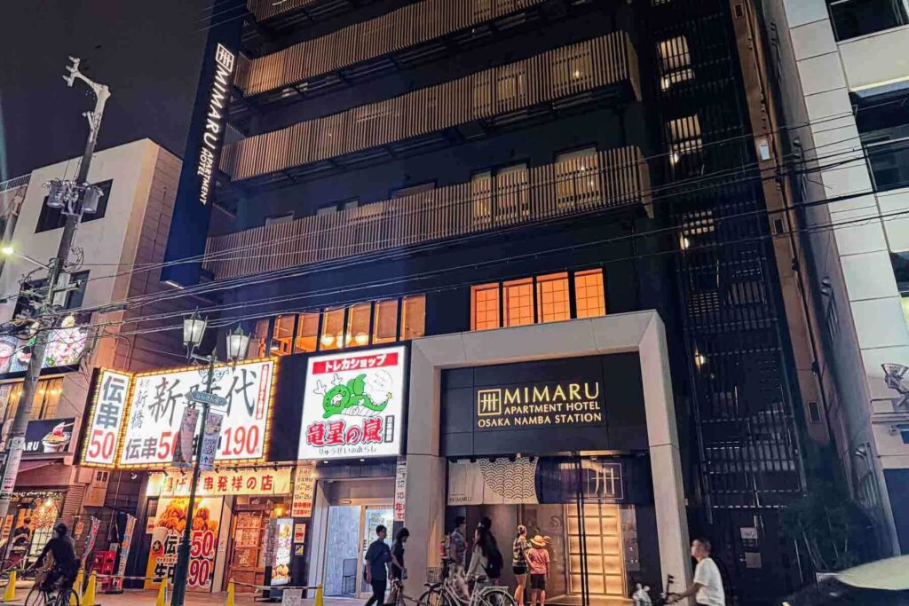 Exterior night view of the Mimaru Osaka Namba Station apartment hotel entrance, illuminated and next to several shops and restaurants.