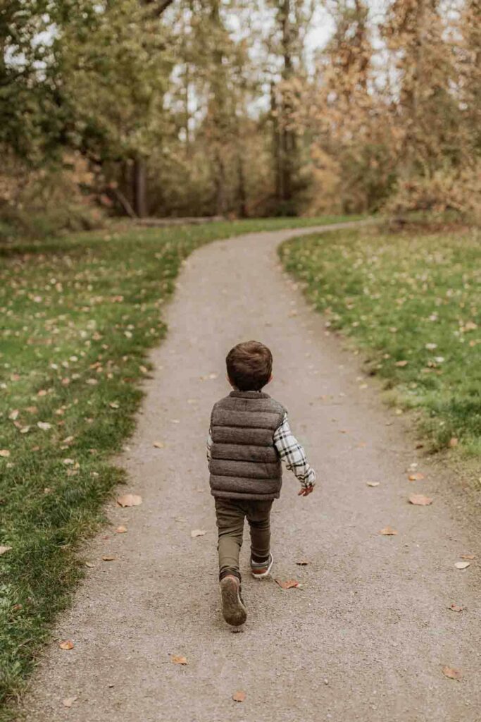 A toddler wearing a gray vest runs away on a paved path with crunchy autumn trees in the background at Washington Park Arboretum.
