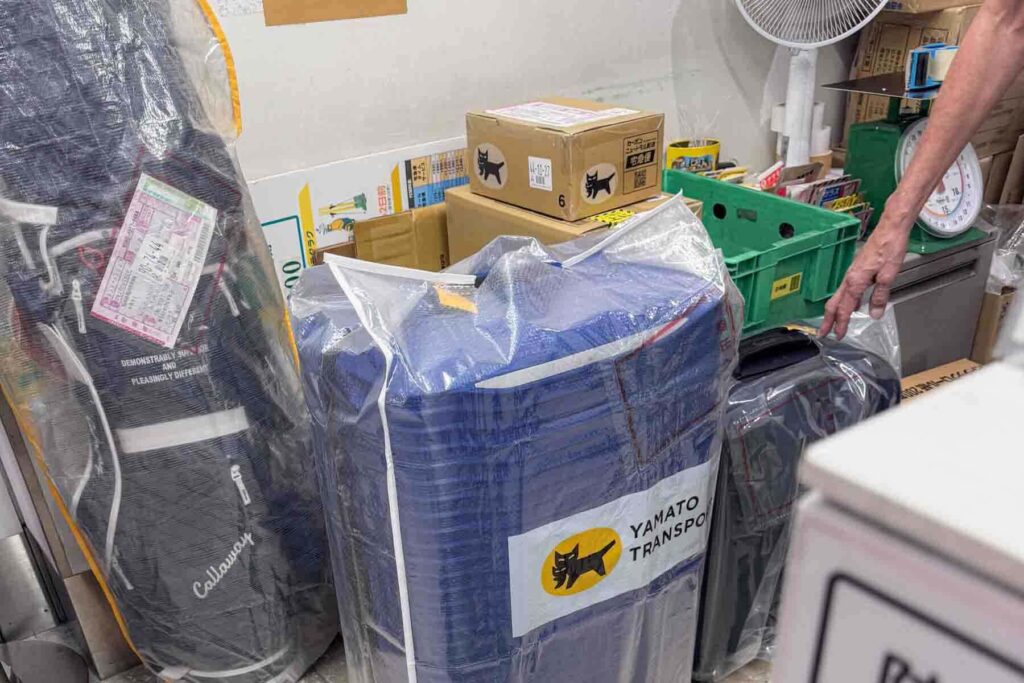 Multiple items, including a suitcase, a golf bag, and cardboard boxes are wrapped and tagged for delivery, all branded with the Yamato Transport logo. Demonstrates how diverse items are safely handled by the takkyubin luggage forwarding service.