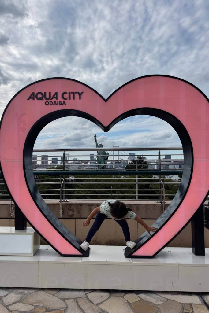 A young child plays inside a giant pink heart sculpture at Aqua City Odaiba, with a view of Tokyo Bay and a replica Statue of Liberty in the background.