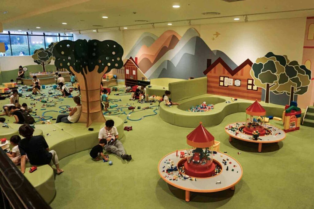 Kids and parents build with blocks, LEGOs and toy trains in a cozy, mountain-themed indoor play area with a padded green floor at Asobono in Tokyo. A perfect indoor play spot for younger kids.