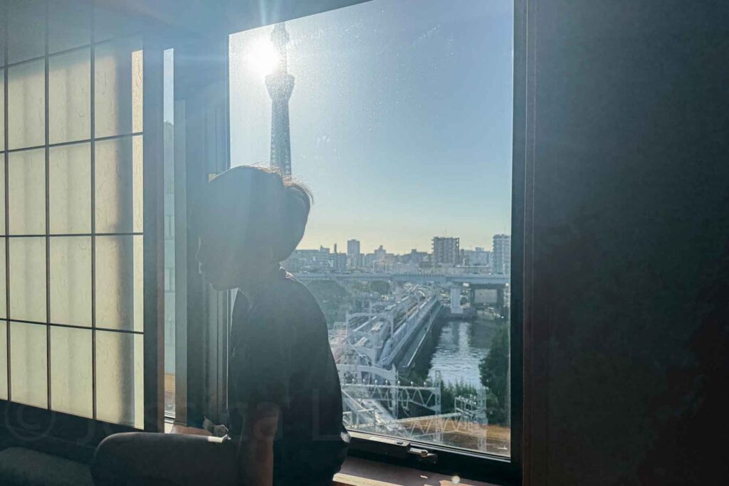 A child sits in silhouette by a large hotel window with a view of Tokyo Skytree, a train track below, and the Sumida River bathed in morning light. Scenic family-friendly moment at Mimaru Suites Tokyo Asakusa.