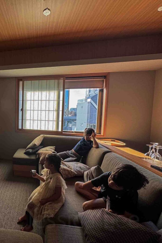 Three children relax and wake up on a large sectional sofa inside a  suite with shoji windows at a modern Japanese family-friendly hotel.