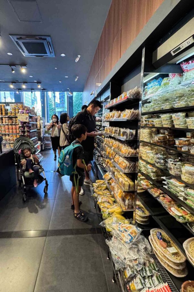 A dad with a young girl in a stroller and boy with a backpack browse snacks and ready-made meals in a Japanese convenience store, Ministop in Tokyo.