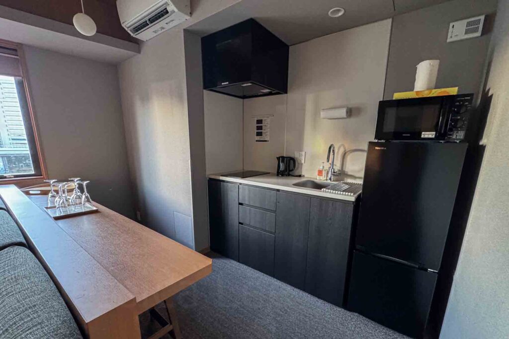 Compact kitchenette with a black fridge and freezer, microwave on top, sink, sleek stovetop, electric tea kettle, and across a wooden dining counter with tray of upside down glasses. Family-friendly amenities in a two-bedroom suite at Mimaru Suites Tokyo Asakusa.