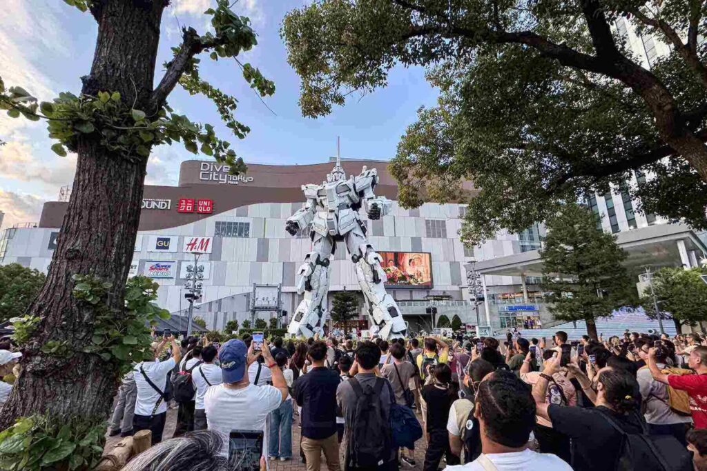 A crowd gathers to admire the giant Unicorn Gundam statue at DiverCity Tokyo, many capturing photos and videos as it moves.