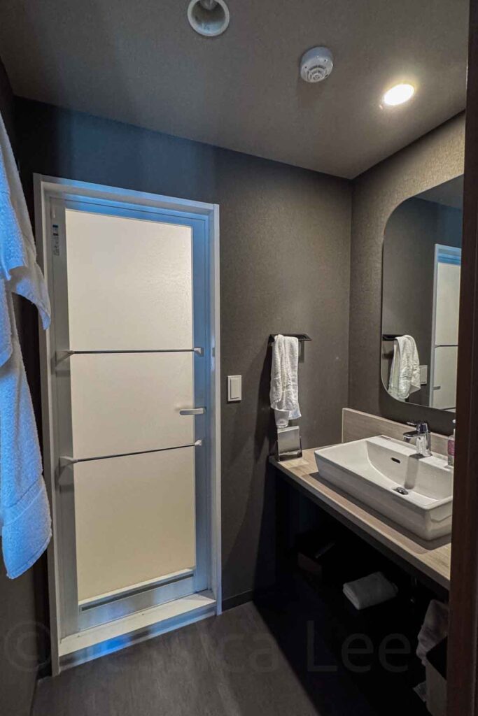 Modern hotel bathroom with dark gray walls a clean white sink vanity, mirror, and door with frosted glass and towel bars leading to the shower room. Convenient and private bath setup with separate facilities great for families staying at Mimaru Suites Asakusa.