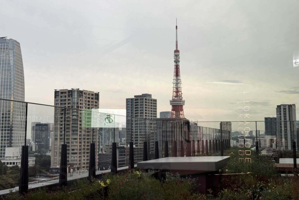 View of Tokyo Tower rising above city skyscrapers from a rooftop observation deck with glass panels.