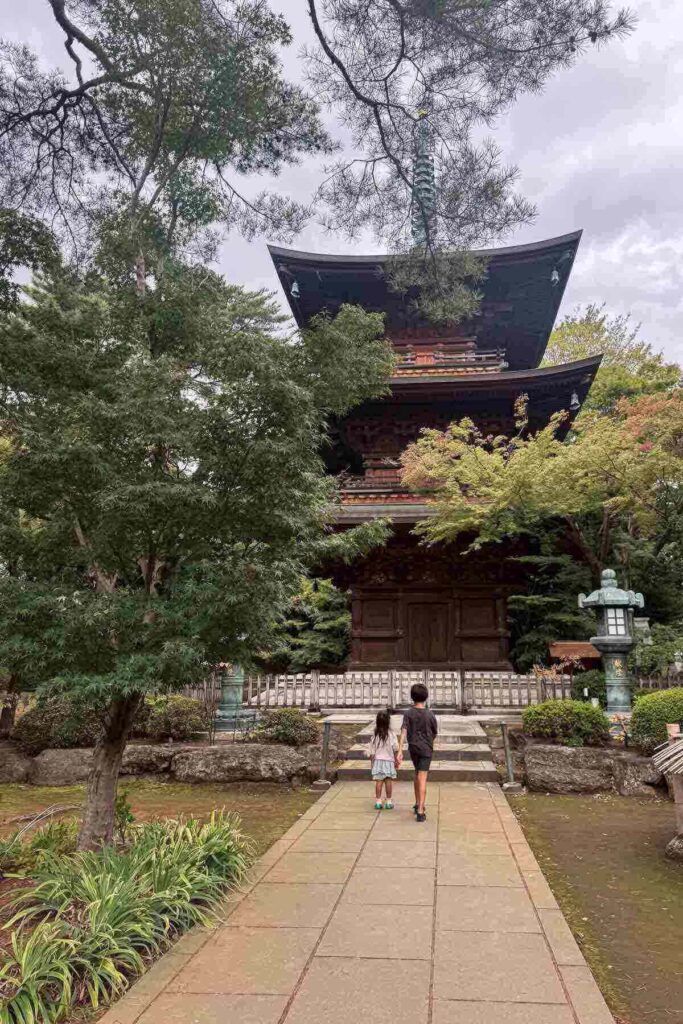A young girl and her brother walk toward a traditional Japanese wooden three story pagoda framed by trees at a quiet Tokyo neighborhood temple, Gotokuji. Exploring peaceful temples is a cultural must and nice break from the bustle of the city.