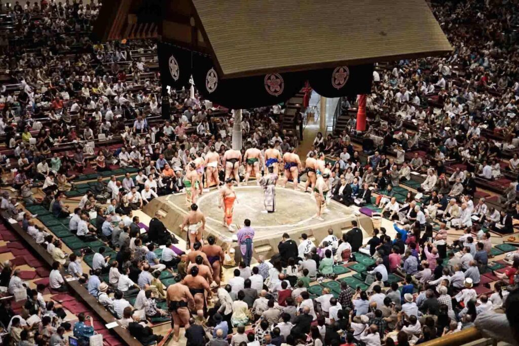 A packed stadium with a large crowd watches sumo wrestlers standing in a circle facing the crowd as they prepare for a match in a traditional ring. Attending a sumo tournament offers a fascinating cultural experience for families exploring Tokyo.