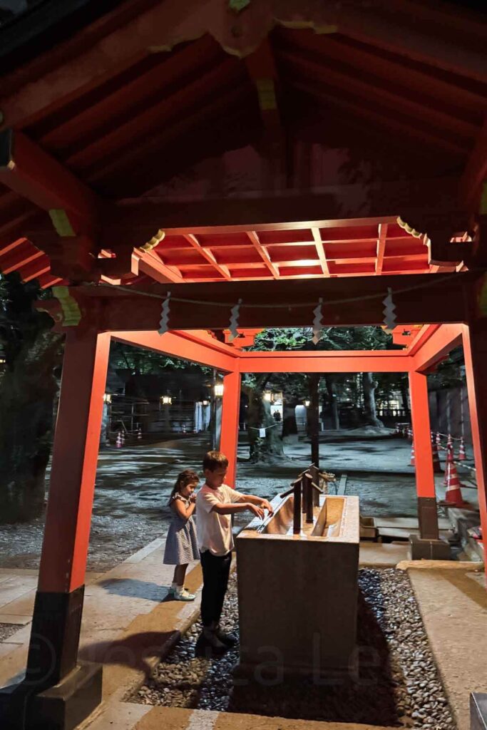 Two children participate in a hand-washing ritual at a red Shinto purification fountain at night the Hanazono Shrine.