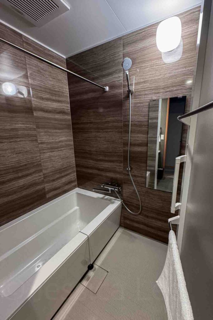 Japanese-style wet bathroom with white deep soaking tub, handheld shower, and brown wood-style wall covering. A long mirror is under the shower and Mimaru provides families with wall-mounted shower toiletries.
