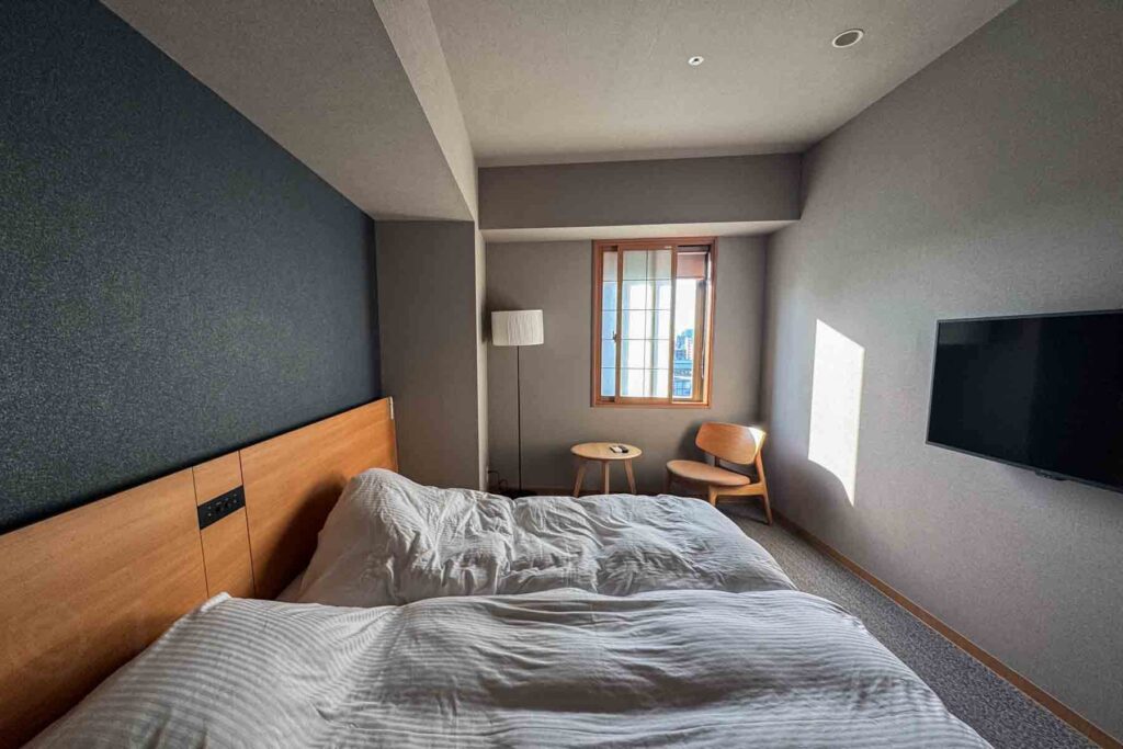 Bright bedroom with dark blue wallpaper accenting one wall with a wooden headboard and two white beds, a wall-mounted TV, and minimalist wooden furniture, a small side table and chair, near a shoji-style window with a simple floor lamp. Peaceful and cozy sleeping area at one of the best places to stay in Tokyo for families.