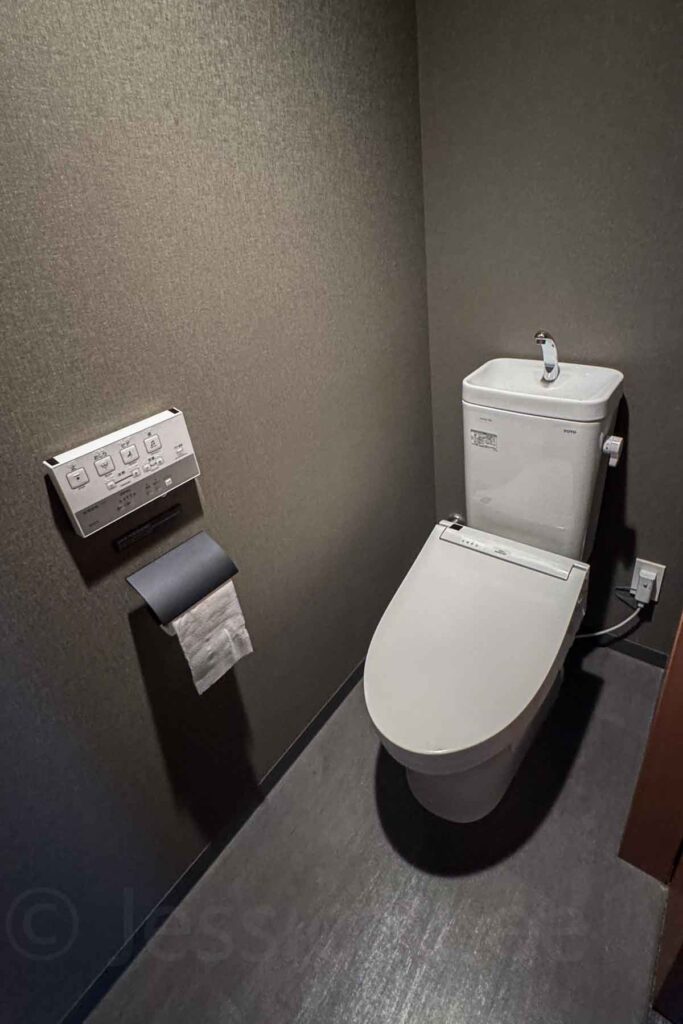 Compact toilet room with dark gray walls featuring a modern Japanese Toto bidet toilet with wall-mounted controls and toilet paper roll. A tiny sink sits on top of the toilet’s water tank.