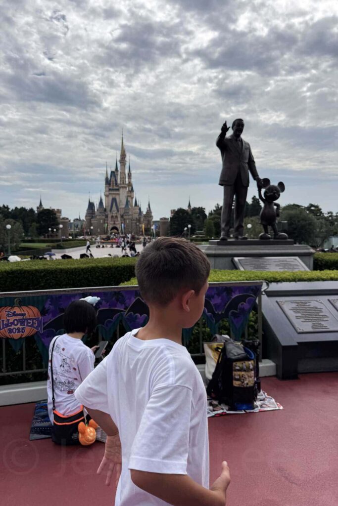 A boy in a white t-shirt stands near the famous statue of Walt Disney and Mickey Mouse at Tokyo Disneyland with Cinderella’s Castle in the distance.