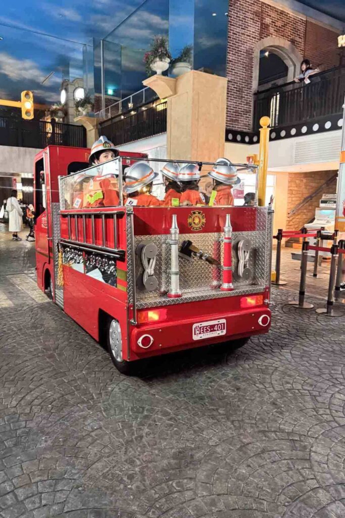 A group of children in orange firefighter uniforms ride the back of a mini fire truck inside KidZania Tokyo’s play pretend city. This immersive role-playing experience is one of the most fun things to do in Tokyo for kids.