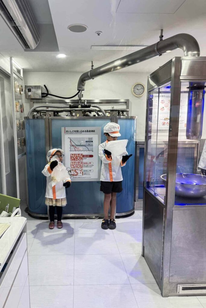 Two kids in white uniforms and hats hold frozen food packs inside a futuristic kitchen lab at KidZania Tokyo. Hands-on activities like this make visiting Tokyo with kids fun and educational.