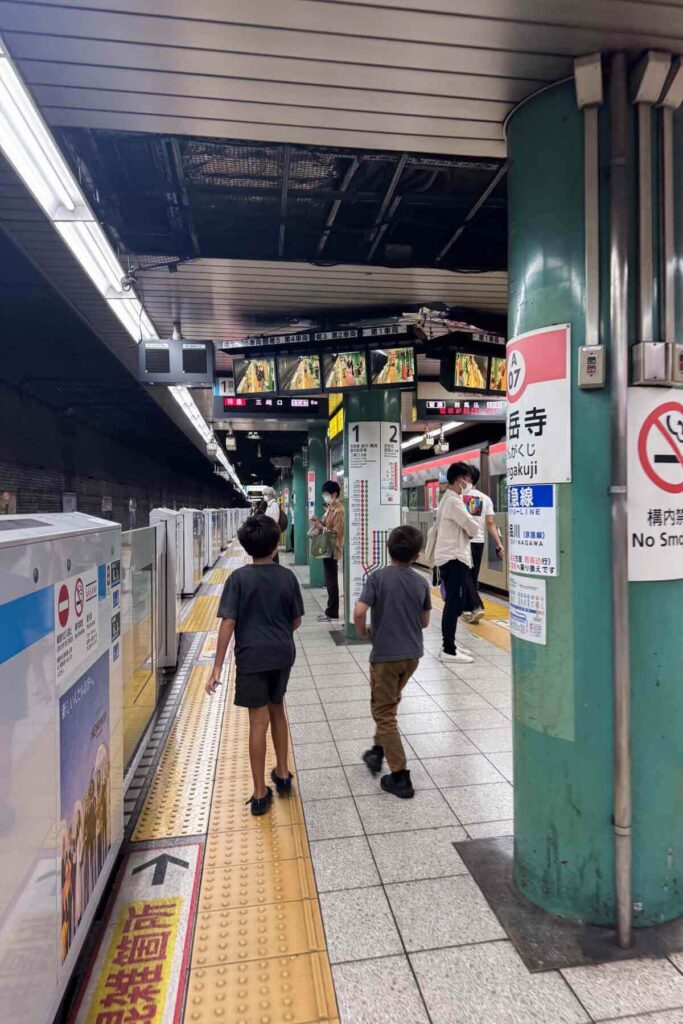 Two elementary-aged boys wait on a Tokyo train platform at Sengakuji Station as an approaching train nears on the opposite track. There are TV monitors above them displaying live video footage of different angles of the station.