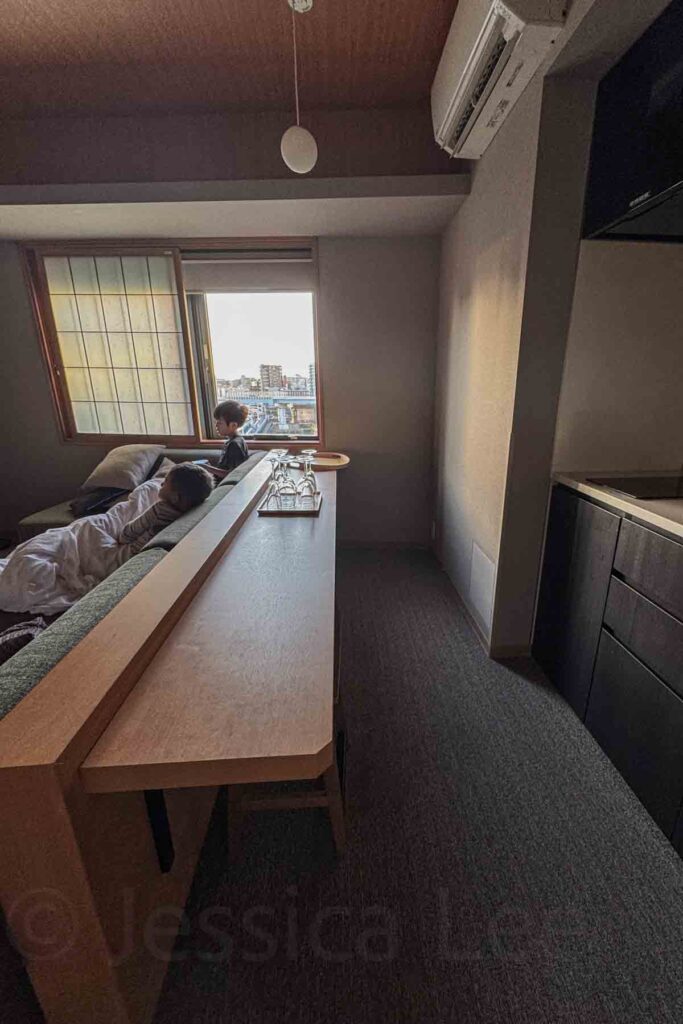 Two children enjoy the cozy living room area in a bright suite with a kitchenette and window city view. Family-friendly layout at one of the best places to stay in Tokyo with kids at Mimaru.