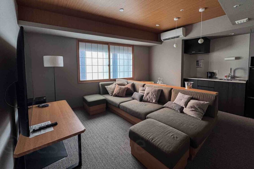 Spacious living room with a large sectional sofa with a wooden frame, shoji windows, and an open black kitchenette. The room has light gray walls and a light wood paneled ceiling. A large TV is at the front of the room with a wooden table below, and a white floor lamp in the corner. Comfortable and modern suite setup at Mimaru Suites Asakusa, perfect for a family of five.