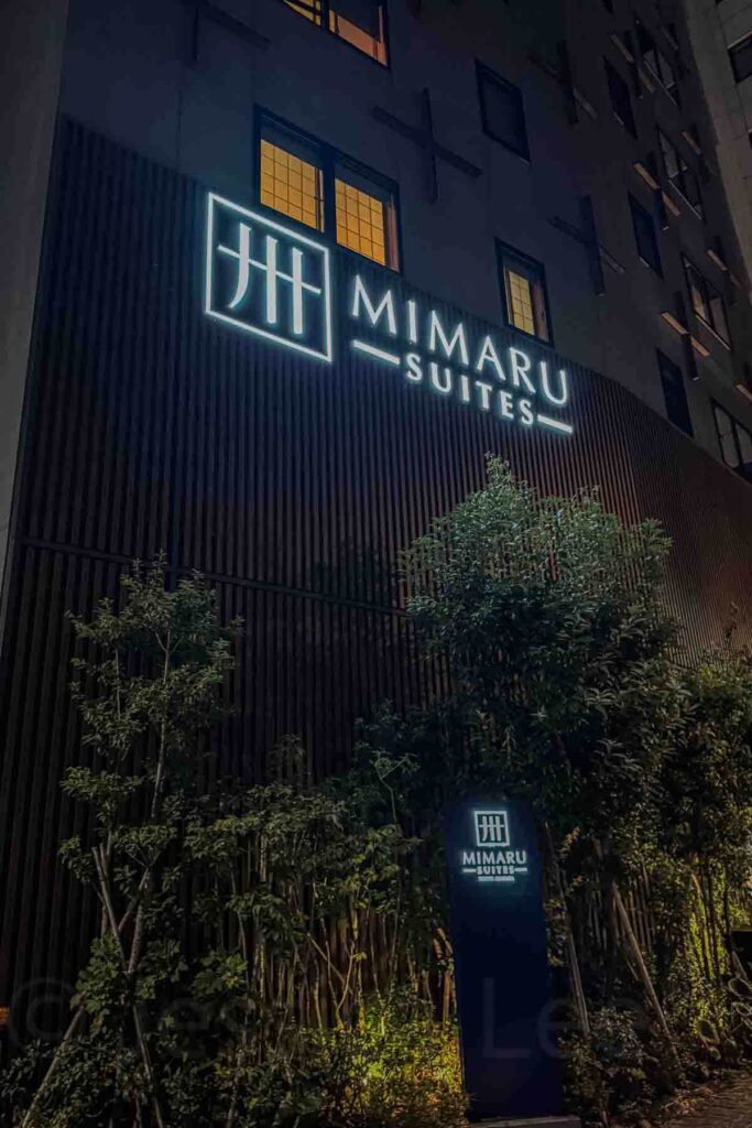 Nighttime view of the modern Mimaru Suites Tokyo Asakusa building with illuminated brand signage and glowing windows, and trees below. 