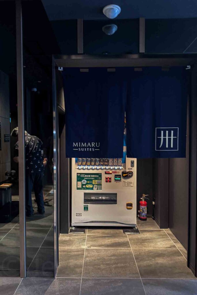 Vending machine corner in the lobby at Mimaru Suites Tokyo Asakusa behind a blue noren curtain with hotel branding. Kid-friendly hotel perk adding convenience for families during their stay.