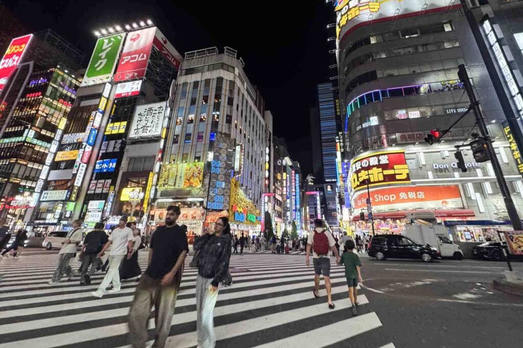 Families and tourists cross an extra wide street in Shinjuku at night, surrounded by bright lights and towering signs.
