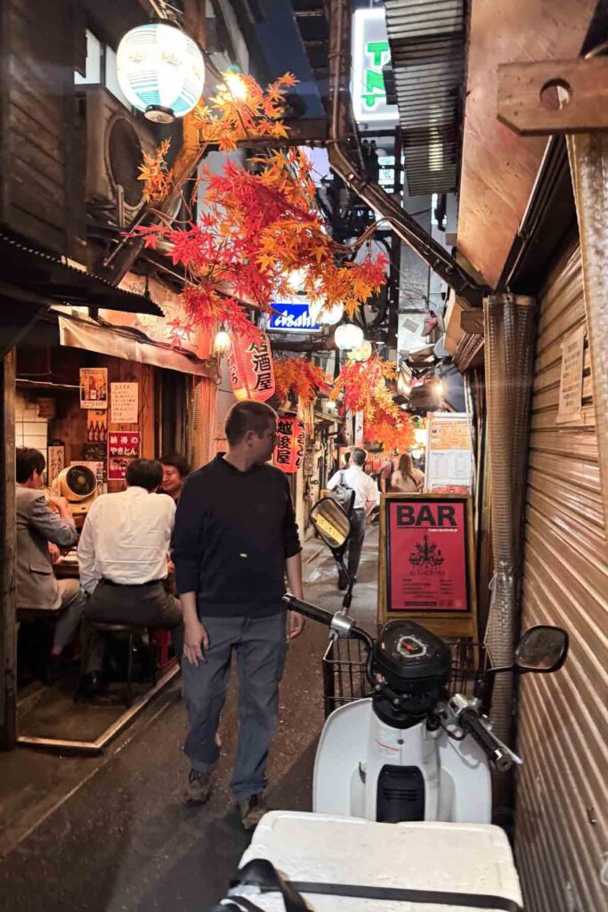 A super narrow alley in Omoide Yokocho in Tokyo filled with lanterns, cozy izakayas, and autumn decorations, as people dine and explore.