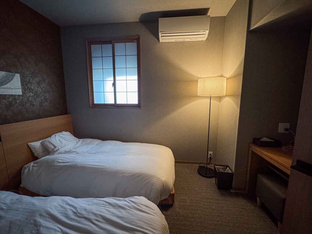 Cozy secondary bedroom with wall mounted wood headboard with two white beds, shoji-style window, desk, and floor lamp. A peaceful sleeping area perfect for families staying at Mimaru Suites Tokyo Asakusa with kids.