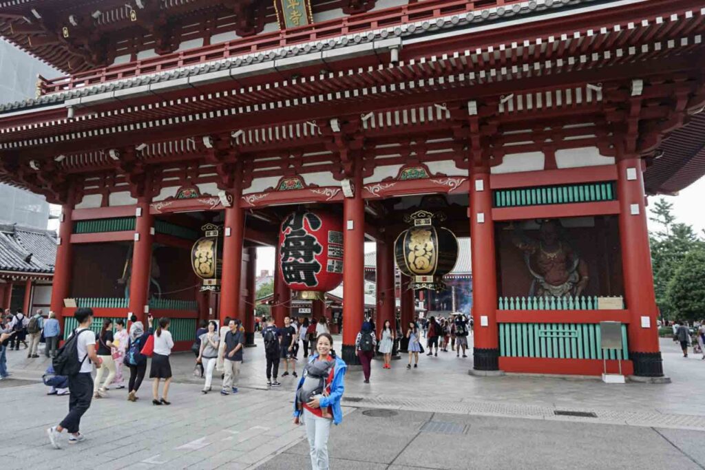 The main gate of Senso-ji Temple bustles with tourists and locals during the day, with a giant red lantern and traditional architecture. A mom stands in front of the gate wearing her baby in a baby carrier. A top cultural experience when visiting Tokyo with kids.