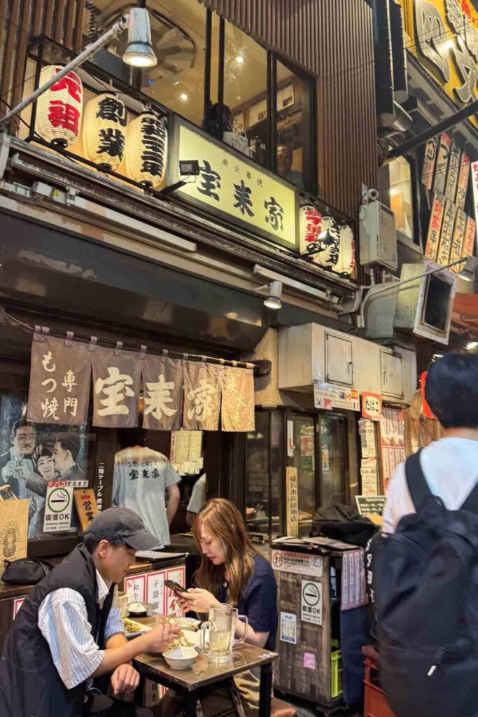 A couple dines at a small outdoor table at a traditional izakaya in Shinjuku, with vintage cloth signage and paper lanterns overhead.