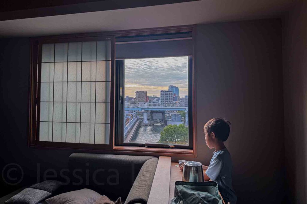 A young boy wearing a blue t-shirt sits at the wooden counter by the window with shoji screen panels overlooking Tokyo's Sumida river and cityscape. Beautiful morning view from a family-friendly suite at Mimaru Suites Asakusa.