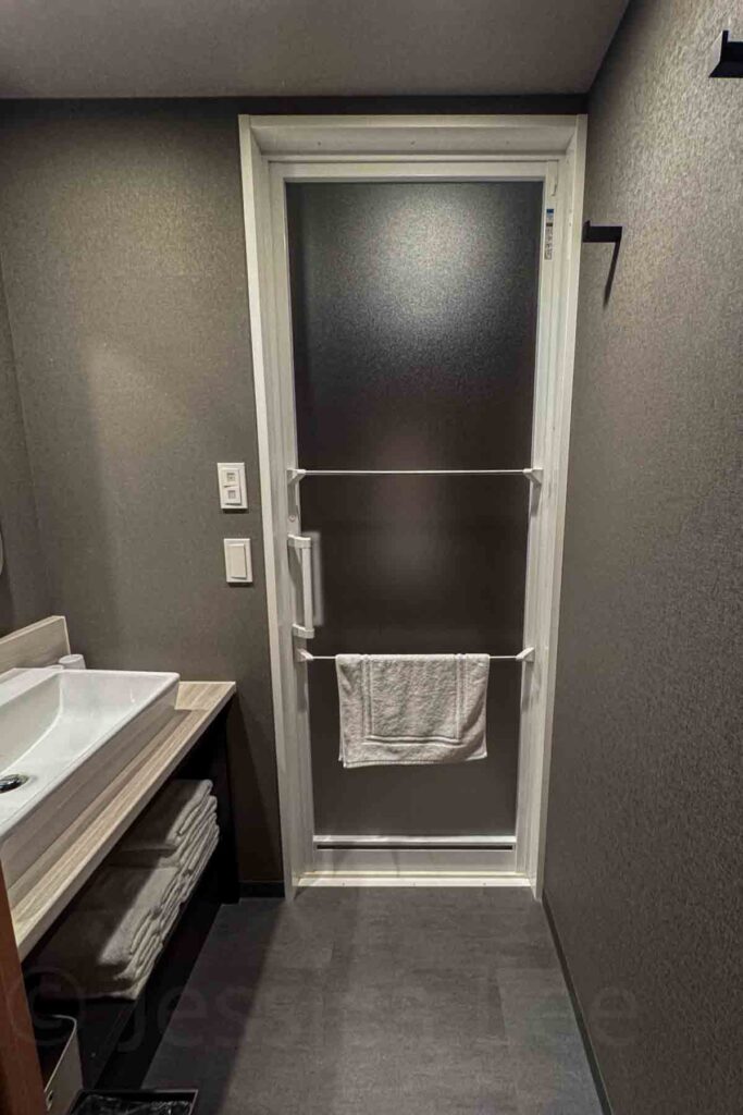Modern Mimaru Suites Tokyo Asakusa hotel bathroom featuring a white vanity sink, stacked towels below, and a frosted glass door with two towel bars leading to the shower. 
