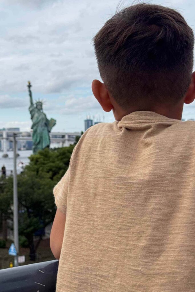 A young boy in a khaki tshirt gazes toward the Statue of Liberty replica in Odaiba, Tokyo Bay stretching behind it under a partly cloudy sky. A scenic and free thing to do in Tokyo with kids.
