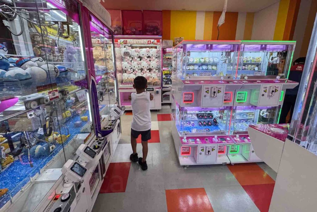 A young boy stands in a brightly lit colorful Taito Station arcade in Shinjuku, Tokyo, surrounded by claw machines filled with plush toys and prizes.