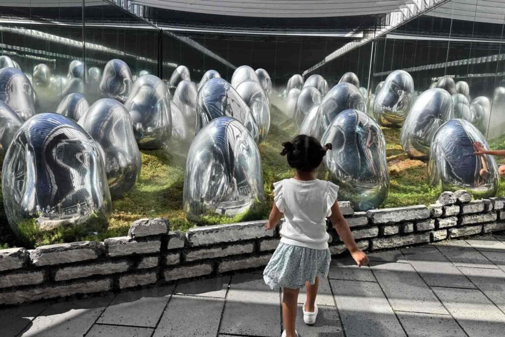 A 3 year old girl walks toward a surreal field of mirrored egg-shaped sculptures at TeamLab Planets Tokyo, with moss, mist and reflective walls creating an engaging effect. This digital art museum captivates kids.