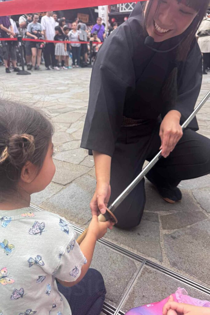 A smiling performer in black ninja outfit lets a young girl hold a samurai sword during a demonstration, as a crowd watches in the Toyosu Market area.