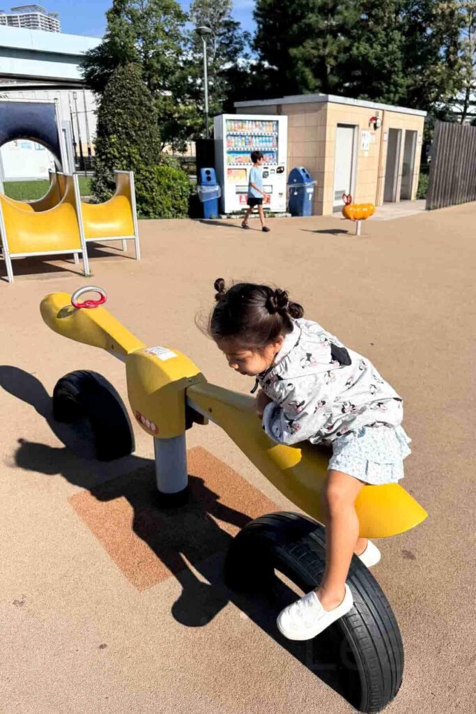 A preschool girl balances on a yellow see-saw at a public Tokyo playground on a sunny day, with vending machines, toilets and slides in the background.