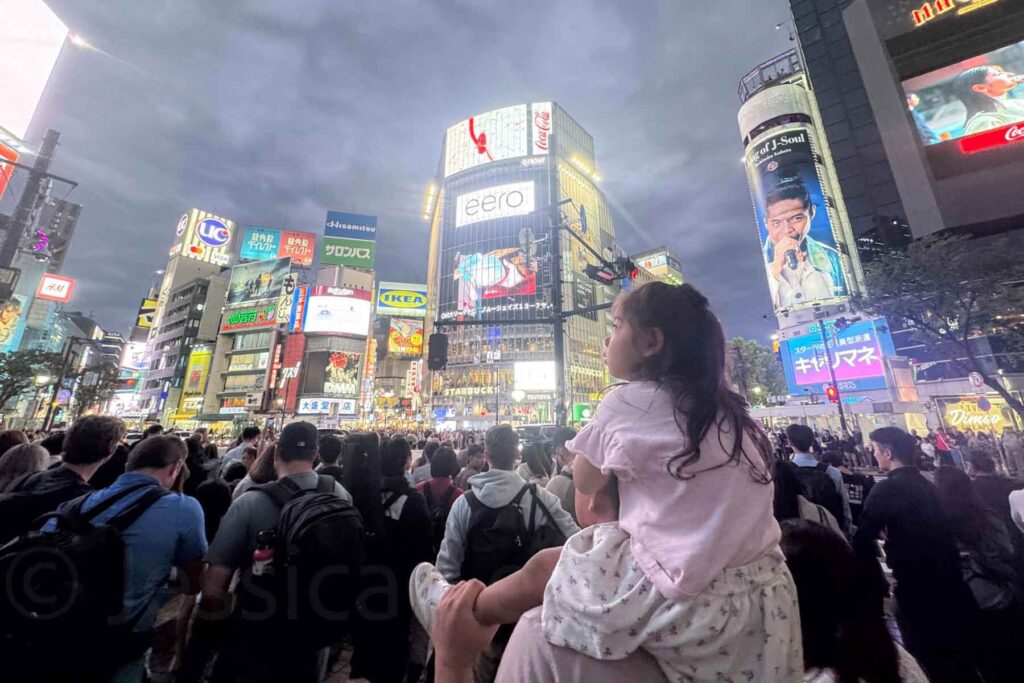 A young girl sits on her dad’s shoulders sitting above a dense crowd while gazing out at the glowing billboards and crowd at Shibuya Crossing. Experiencing this iconic spot is a thrilling Tokyo moment.